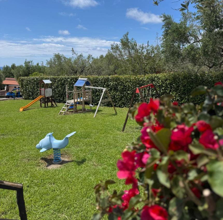 Giardino con giochi per bambini, cielo azzurro, fiori rossi vibranti, atmosfera serena.