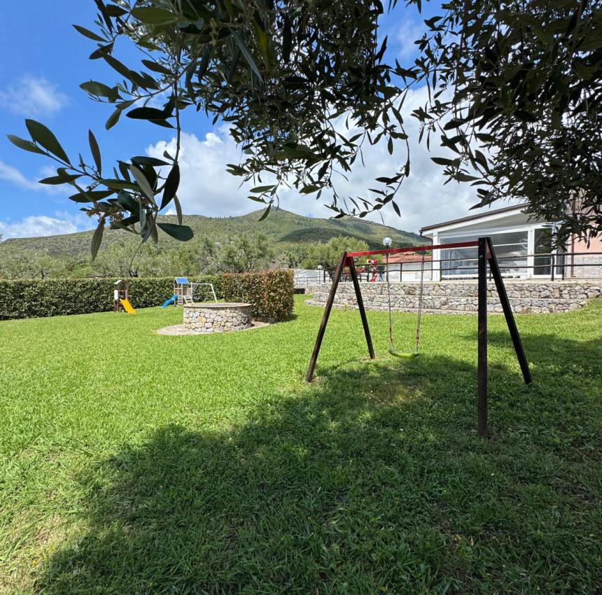 Giardino assolato con altalena, cielo azzurro e colline verdi.
